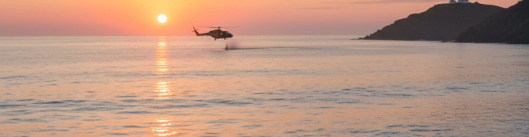 Helicopter flying over ocean waves during a vibrant sunset near a distant lighthouse.