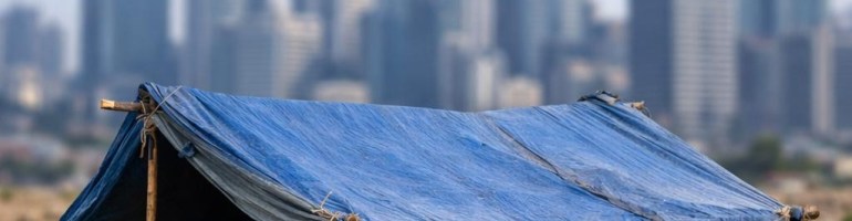 Makeshift blue tarp shelter on dry land with modern city skyscrapers in the background