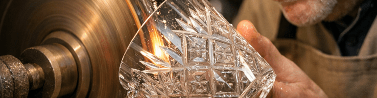 Craftsman engraving intricate patterns on a crystal glass with a rotating grinding wheel and magnifying goggles