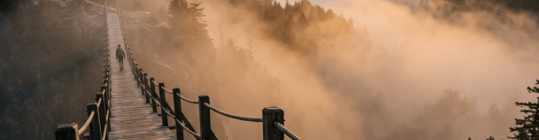 Wooden suspension bridge over foggy valley with a person walking, surrounded by mountains and trees at sunrise.