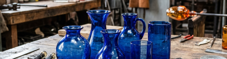 Six blue glass vessels of various shapes on a wooden workbench with glassblowing tools and furnace in background