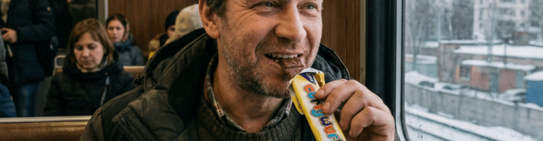 Man eating chocolate bar and holding thermos on train seat by window showing snowy urban landscape