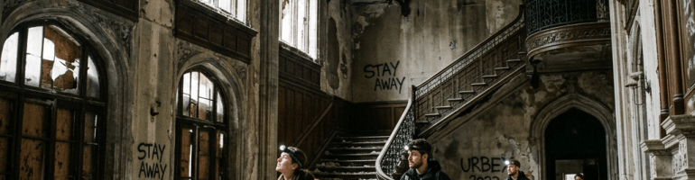 Four people walking inside a dilapidated gothic-style building with broken windows and graffiti saying 'STAY AWAY' and 'URBE 2023'