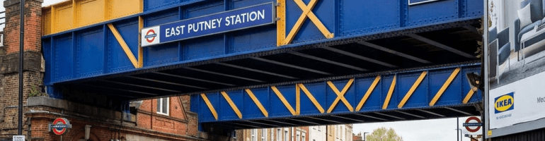 Railway bridge painted blue and yellow with East Putney Station sign over a busy London street