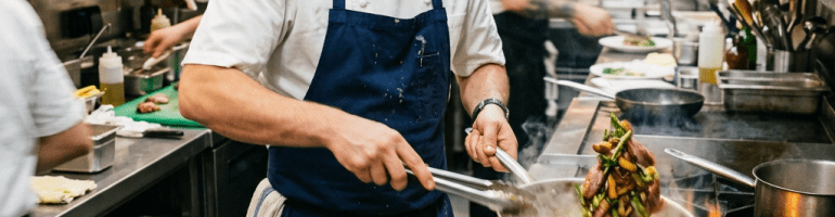 Chef cooking vegetables and meat in a flaming pan in a busy professional kitchen