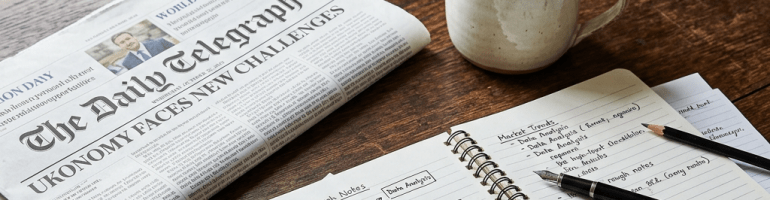 Newspaper titled The Daily Telegraph, open notebook with notes and pens, steaming cup of tea, small potted plant, stack of books on wooden table