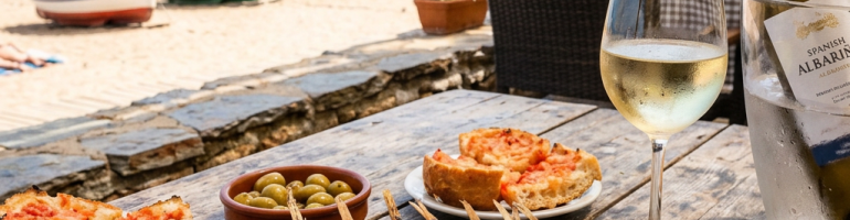 Grilled sardines on skewers with lemon wedges on a plate, bread with tomato, olives, and a glass of white wine by the seaside