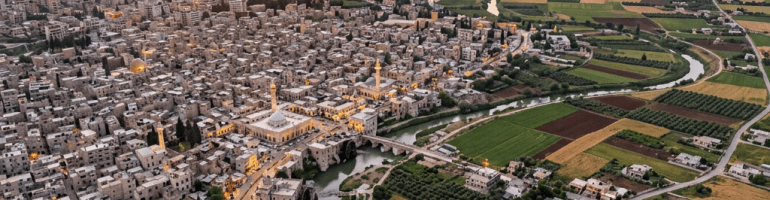 Aerial view of a town with clustered buildings near a winding river, surrounded by green and brown farmland at sunset.