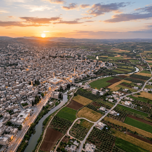 Aerial view of a town with clustered buildings near a winding river, surrounded by green and brown farmland at sunset.